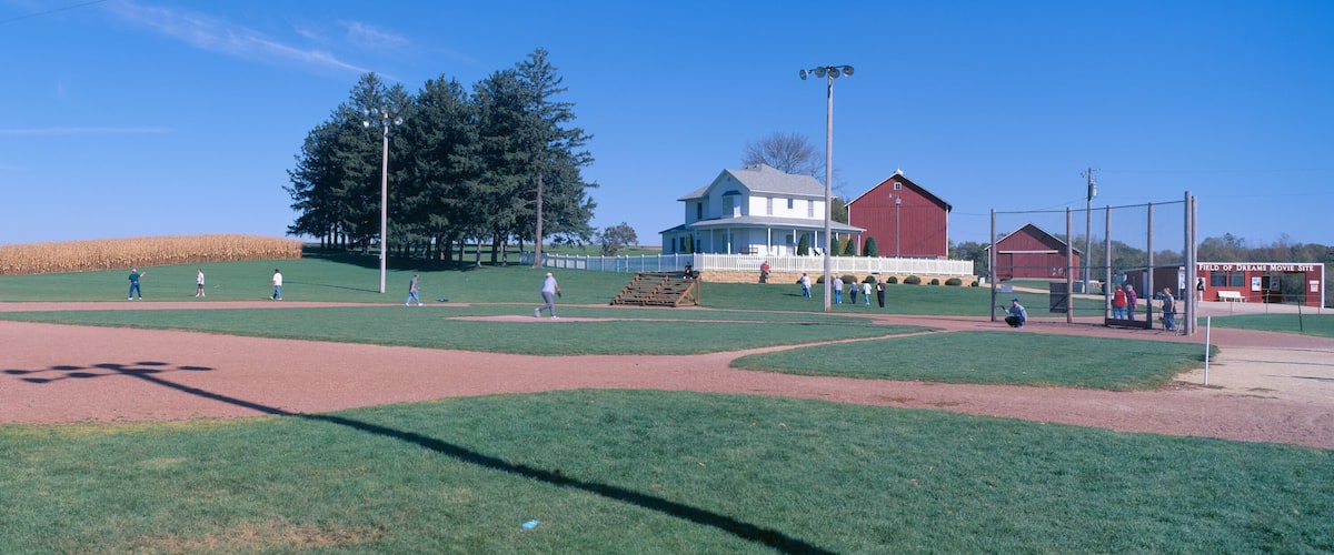 A baseball field with a red barn and a white fence in the background.