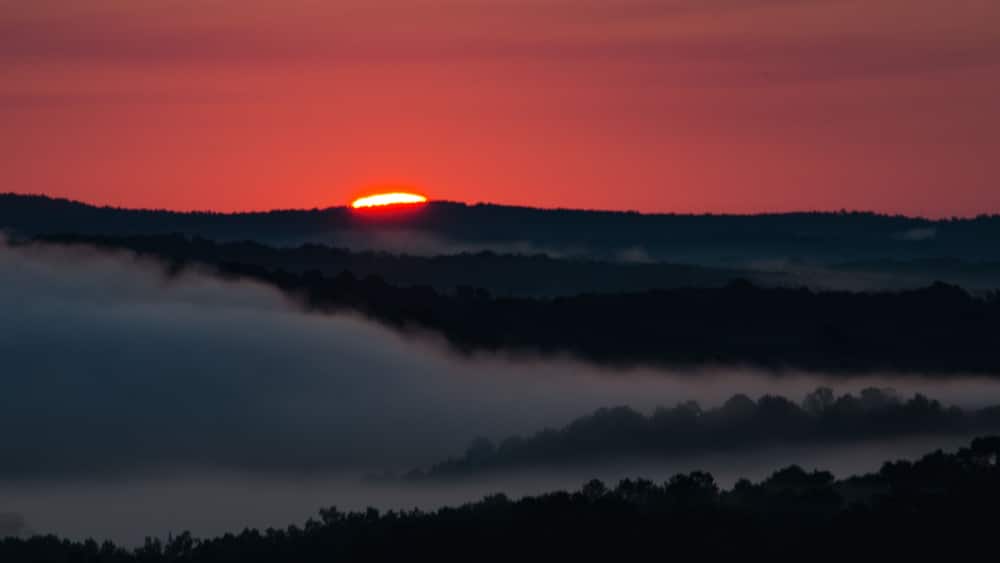 Un coucher de soleil sur un paysage boisé, le soleil étant partiellement caché par les nuages.