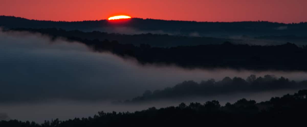 A sunset over a forested landscape with a sun partially obscured by clouds.