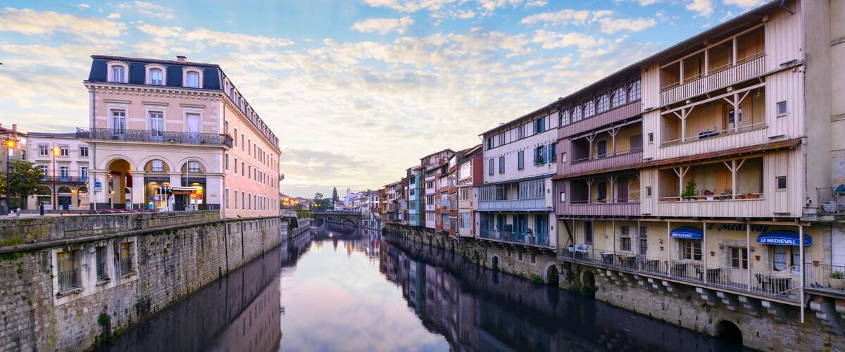 A calm river reflecting buildings, a stone bridge, and a clear sky with clouds.