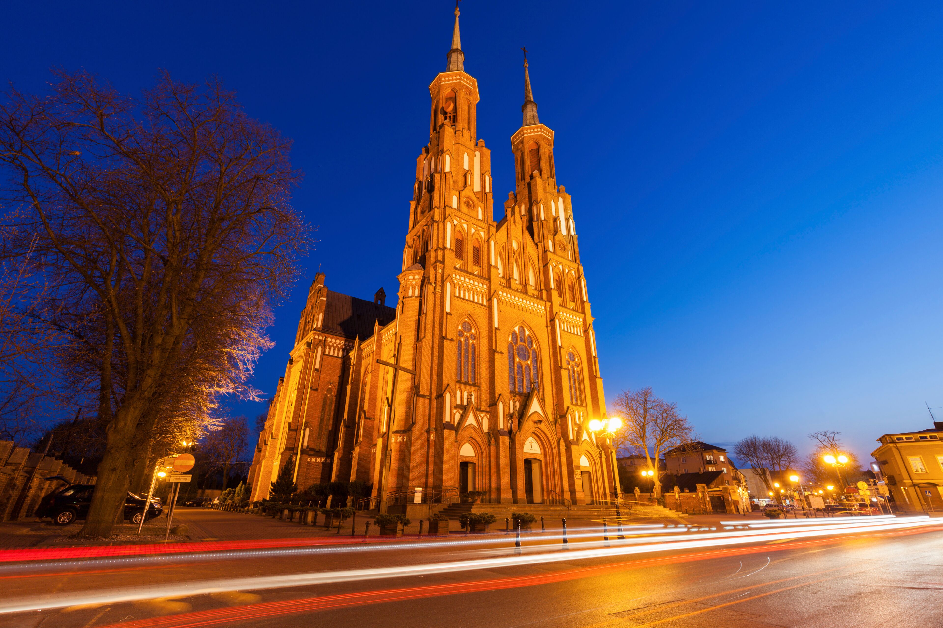A large, illuminated cathedral with twin spires, surrounded by trees and a city street with light trails.