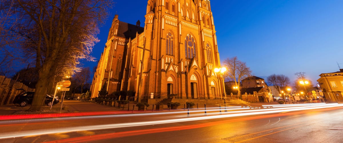 A large, illuminated cathedral with twin spires, surrounded by trees and a city street with light trails.