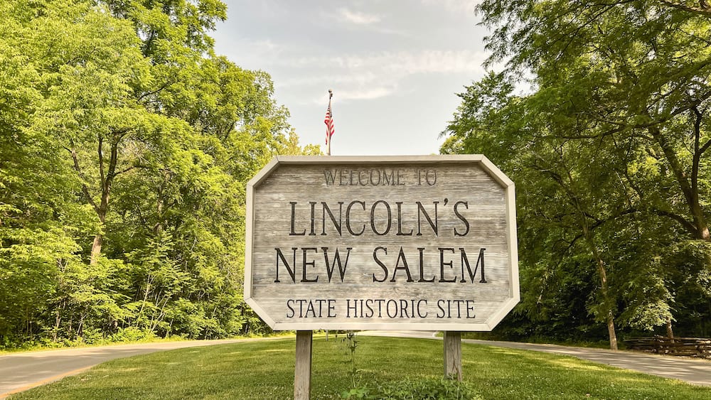 A wooden welcome sign for Lincoln's New Salem State Historic Site surrounded by lush greenery and a clear sky.