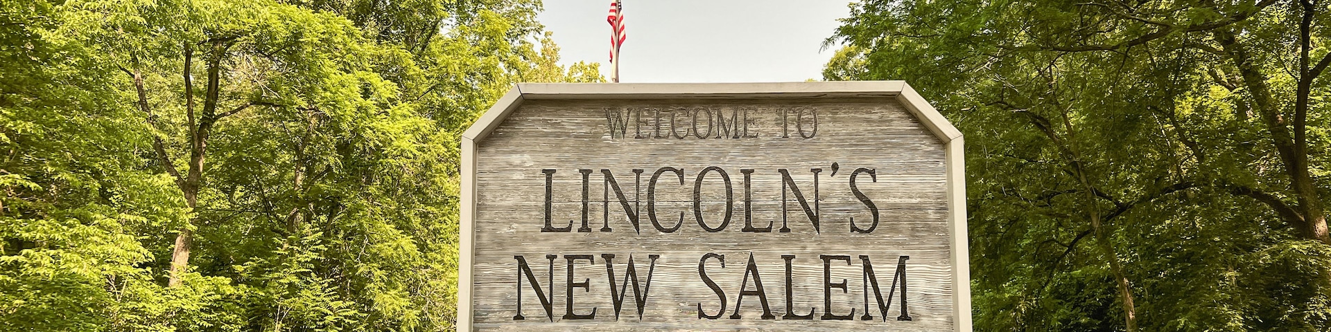 Un letrero de madera da la bienvenida al Sitio Histórico Estatal de Lincoln’s New Salem, rodeado de exuberante follaje y un cielo despejado.