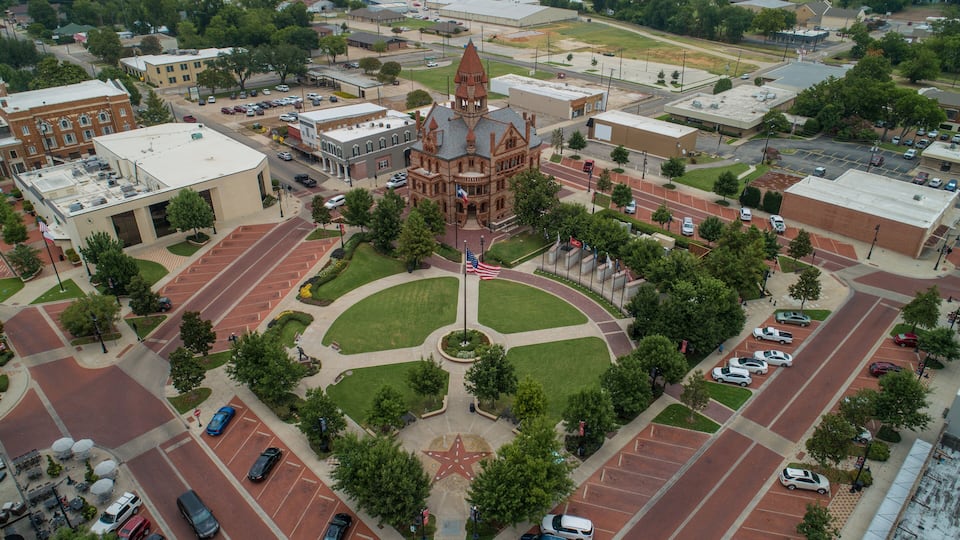 An aerial view of a town square with a prominent historic building, parking lots, and surrounding greenery.