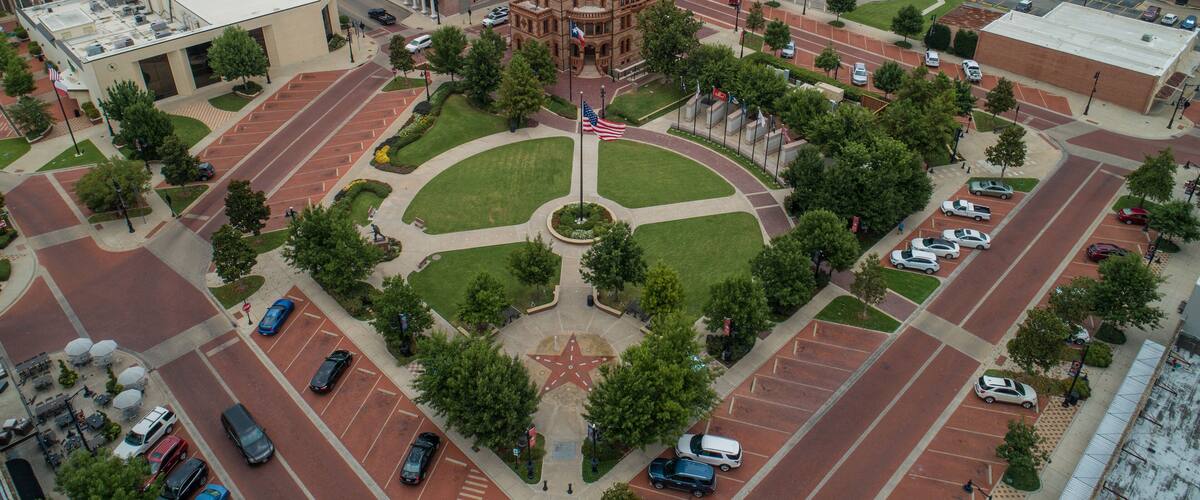 An aerial view of a town square with a prominent historic building, parking lots, and surrounding greenery.