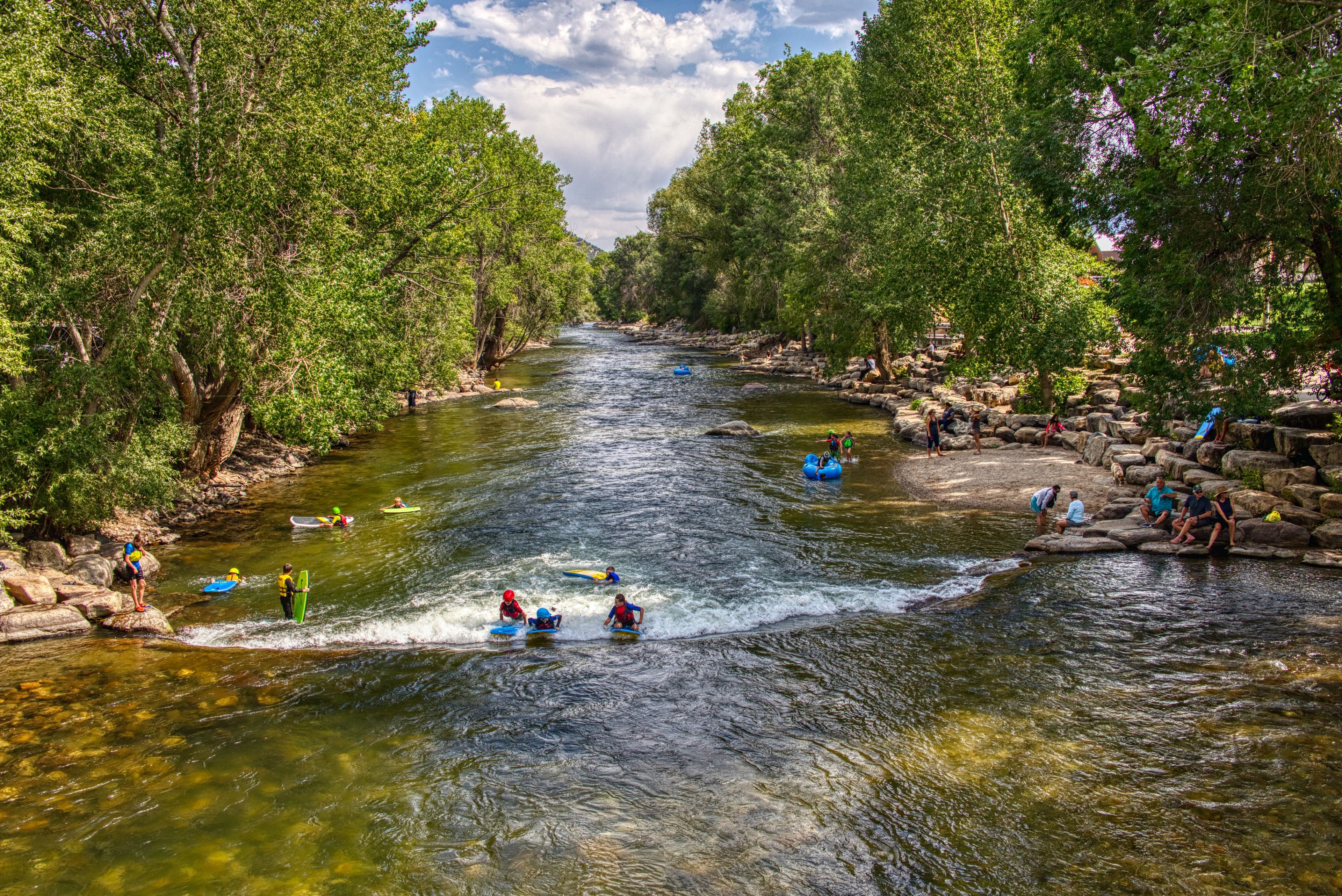 A river surrounded by dense trees, with people kayaking and standing on rocks.