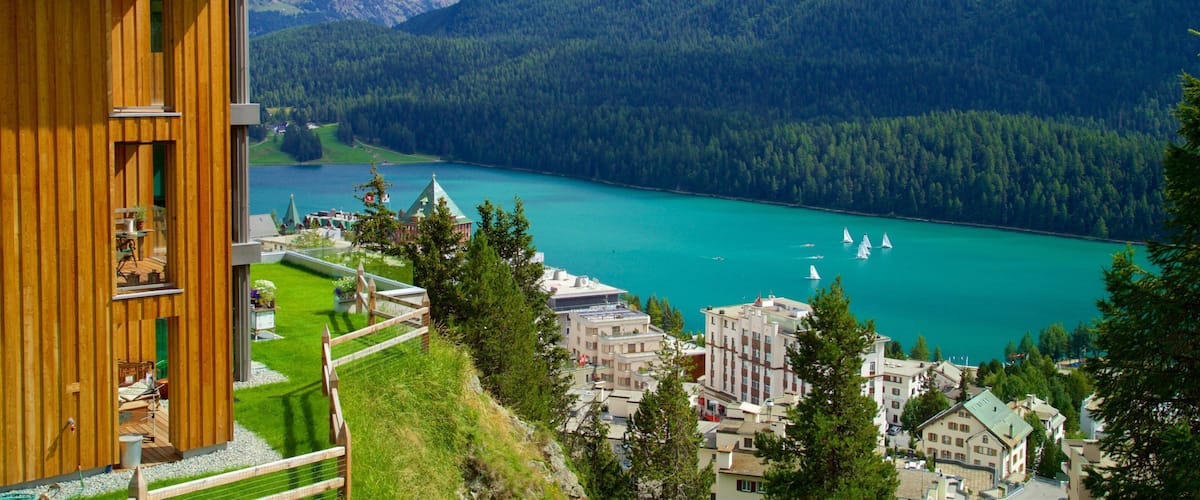 A wooden building overlooking a turquoise lake with sailboats, surrounded by forested mountains.