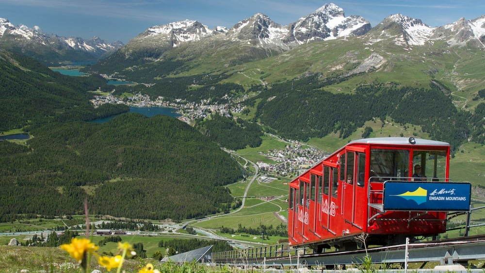 Eine rote Gondelbahn in den Engadiner Bergen, im Hintergrund ein See und schneebedeckte Gipfel.
