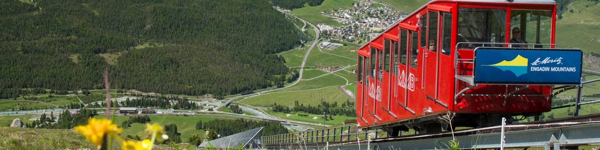 A red cable car in the Engadin Mountains with a lake and snow-capped mountains in the background.