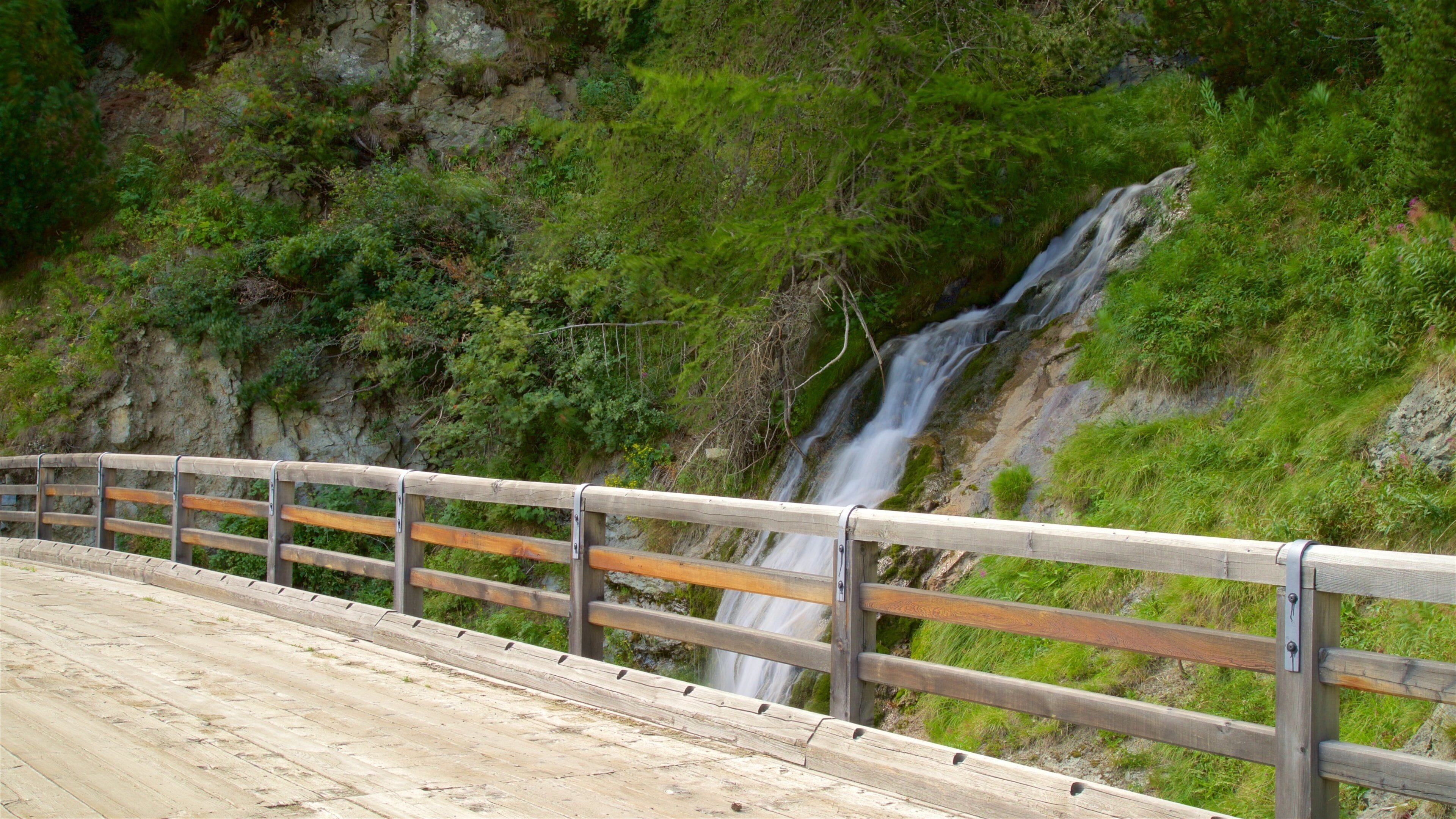 A wooden bridge with a railing, leading to a waterfall cascading down a rocky cliff.