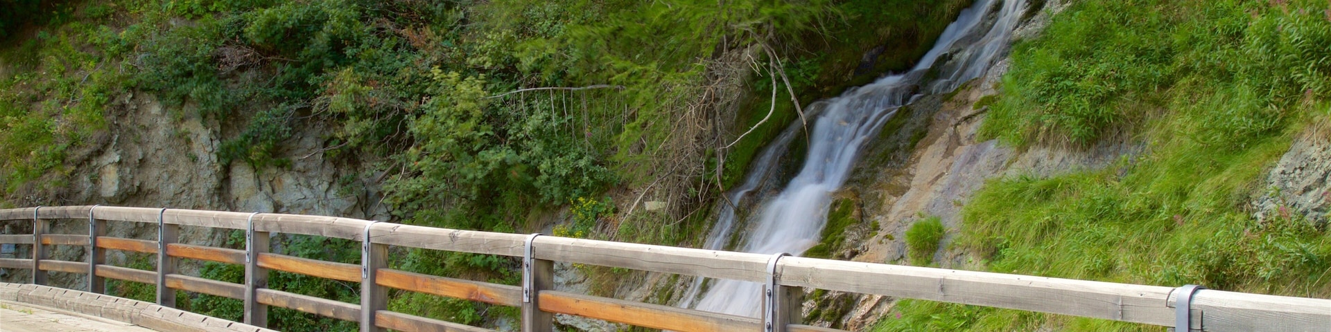 A wooden bridge with a railing, leading to a waterfall cascading down a rocky cliff.