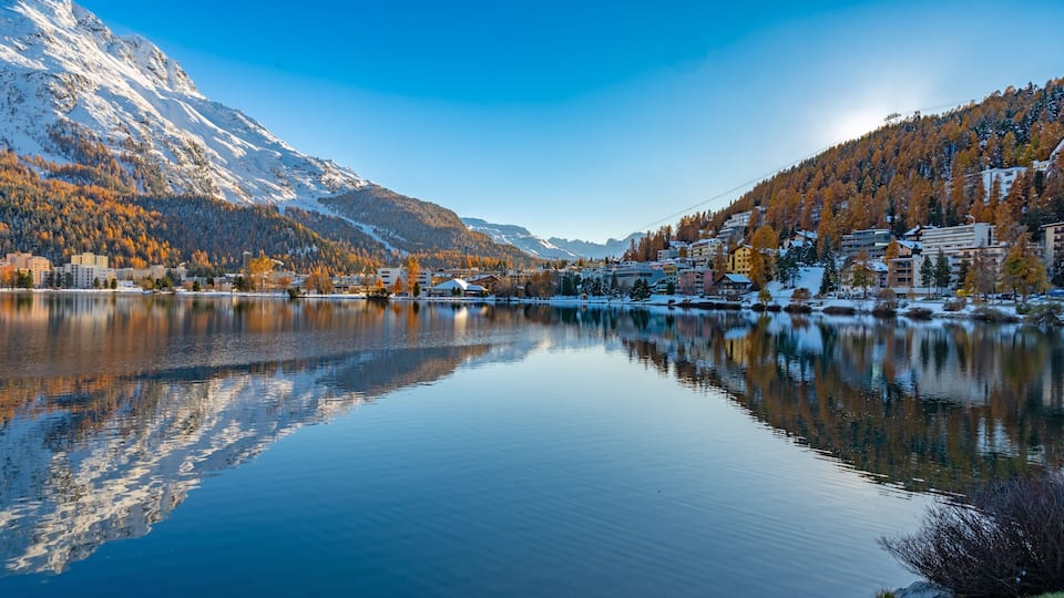 A calm lake reflecting mountains, a town with buildings, and autumn-colored trees.