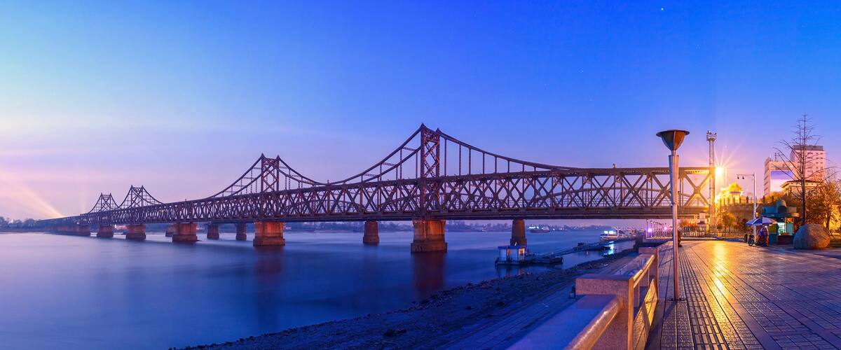 A large steel bridge spanning a river at dusk with city lights in the background.