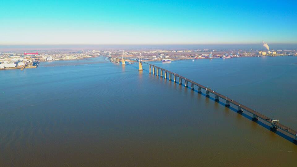 A long bridge spanning a wide river, with industrial buildings and a distant city skyline.