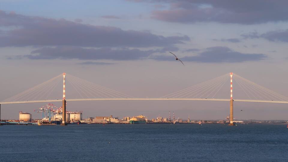 A large suspension bridge with two tall towers and cables, a body of water, industrial buildings, and a ship.