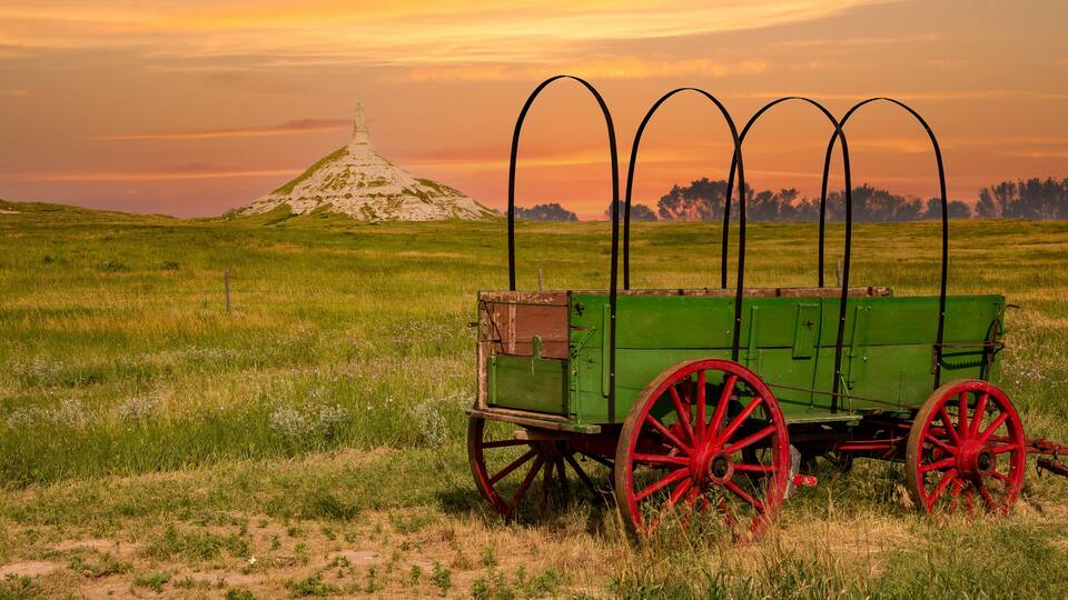 A green cart with red wheels in a grassy field with a hill and a monument in the background.