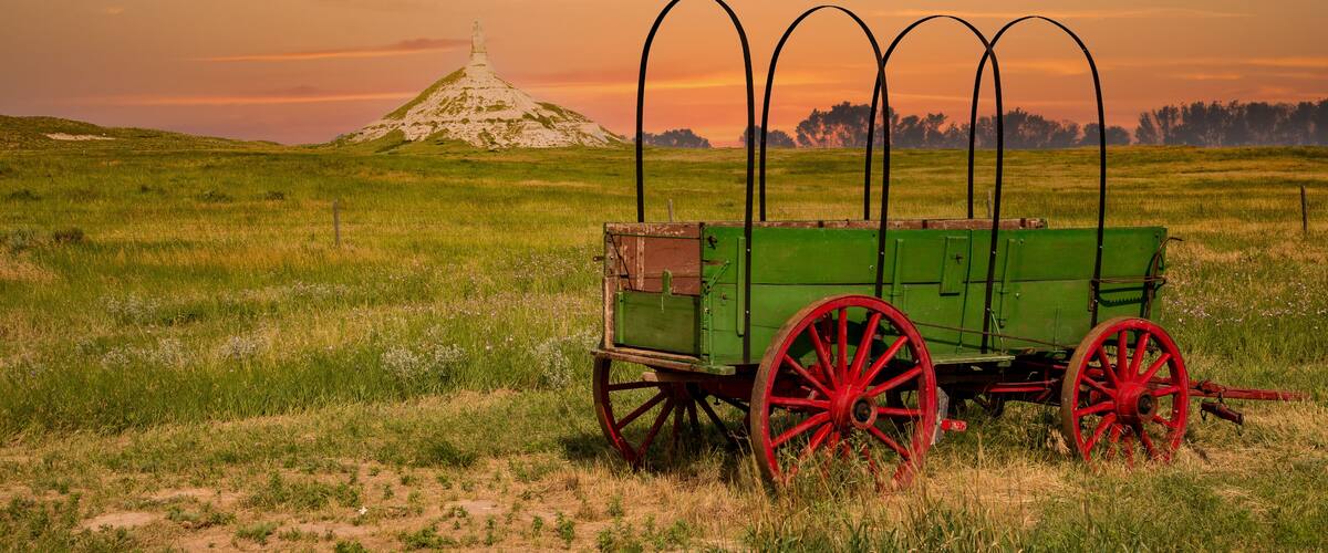 A green cart with red wheels in a grassy field with a hill and a monument in the background.