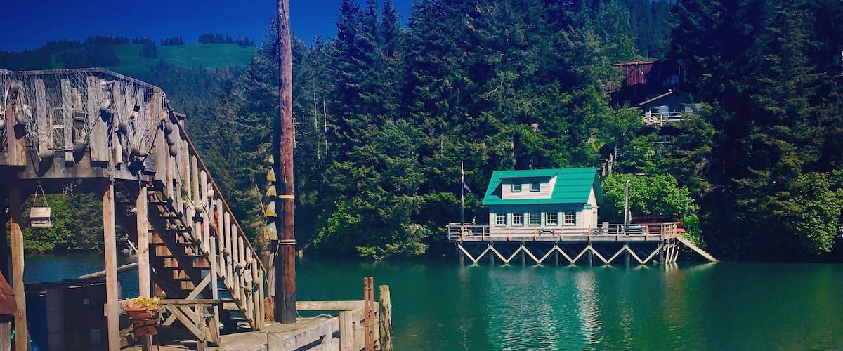 Un muelle de madera con una escalera rústica, una pequeña casa con techo verde y un lago tranquilo rodeado de bosque denso.