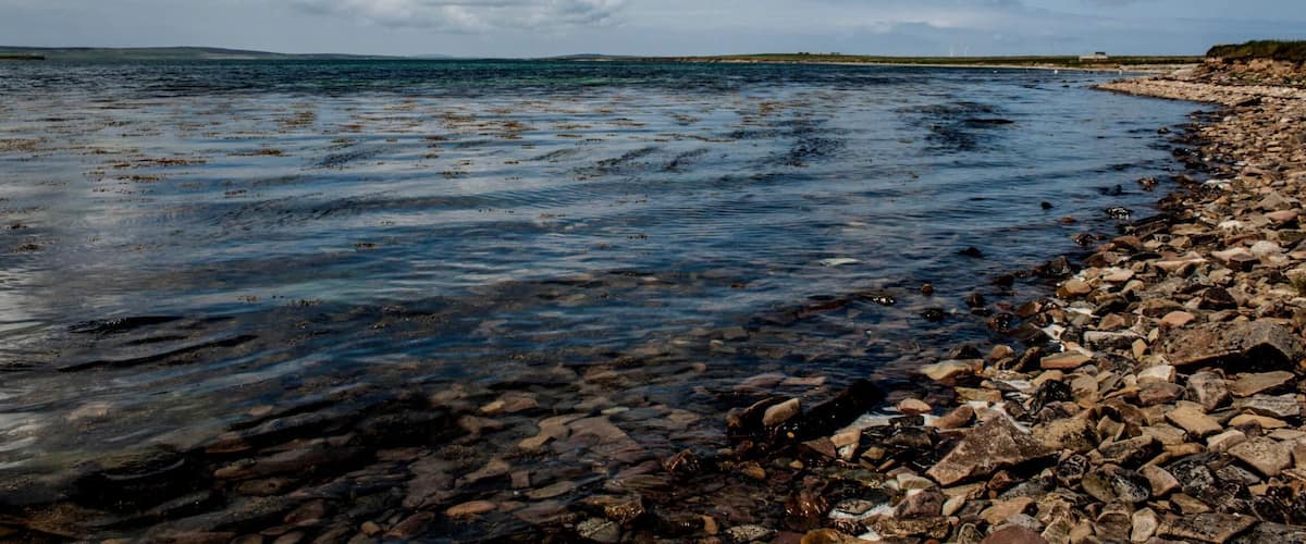 A rocky shoreline with clear water and distant land under a partly cloudy sky.