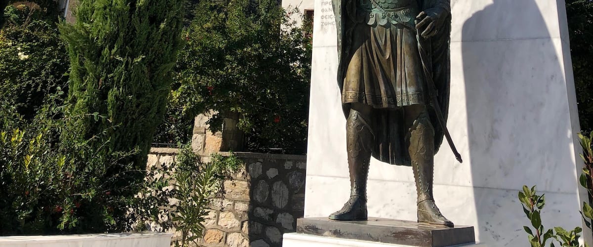 A statue of a historical figure with a flag, set against a mountainous backdrop and a clear blue sky.