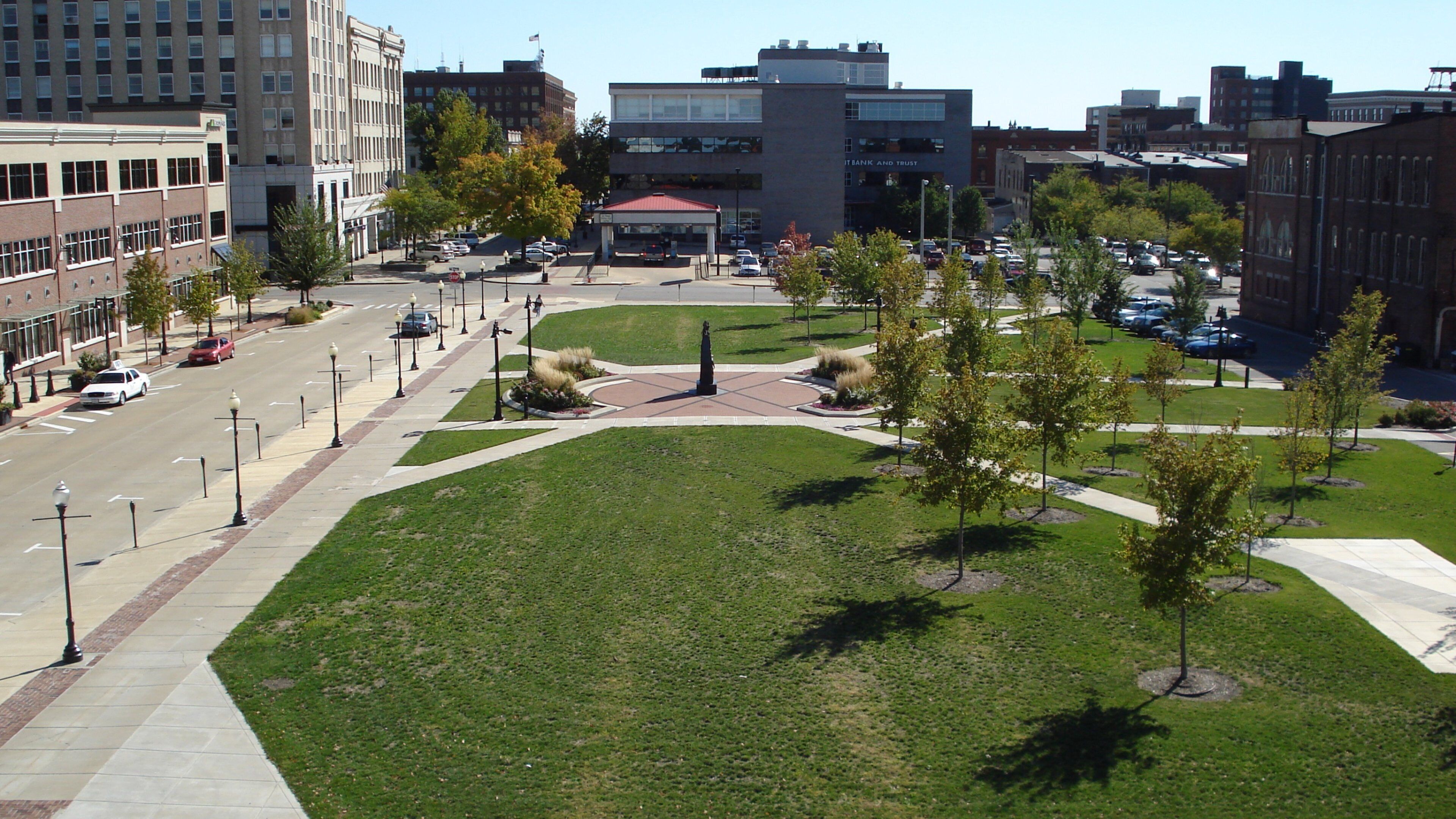 A city square with a central monument, surrounding buildings, and a parking lot.