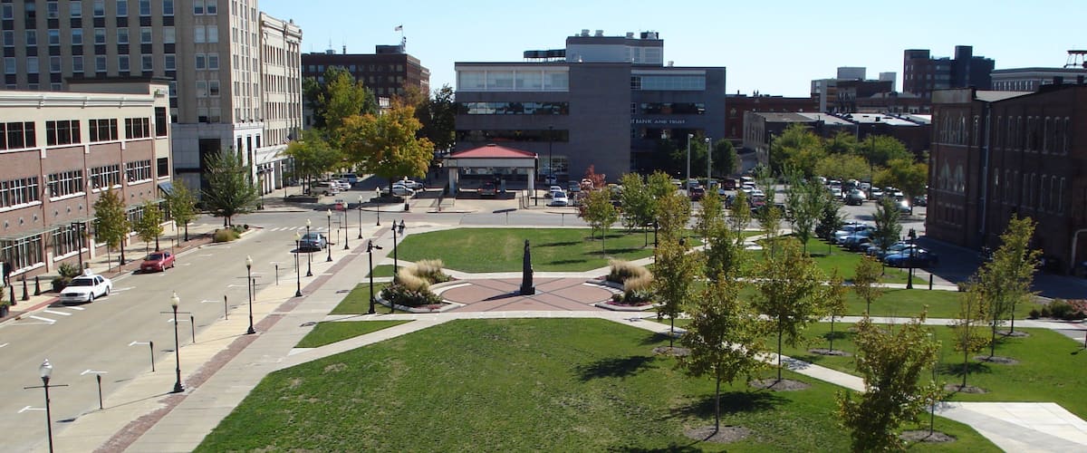 A city square with a central monument, surrounding buildings, and a parking lot.