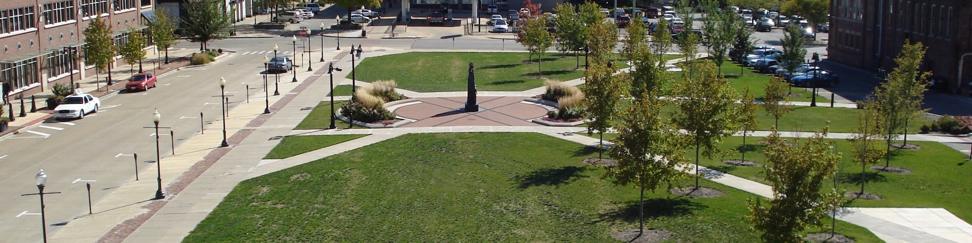 A city square with a central monument, surrounding buildings, and a parking lot.