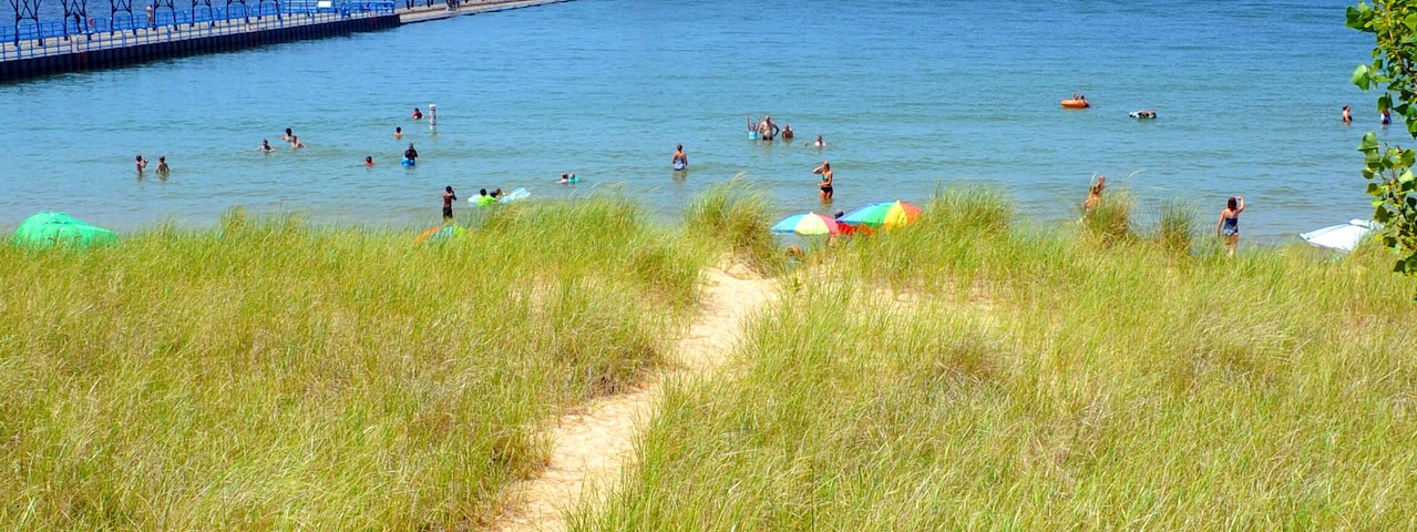 Un camino costero que lleva a un faro, gente en la playa y un muelle que se adentra en el mar.
