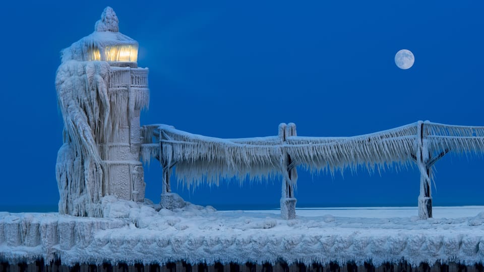 A frozen bridge with icicles, a lighthouse, and a full moon in the night sky.