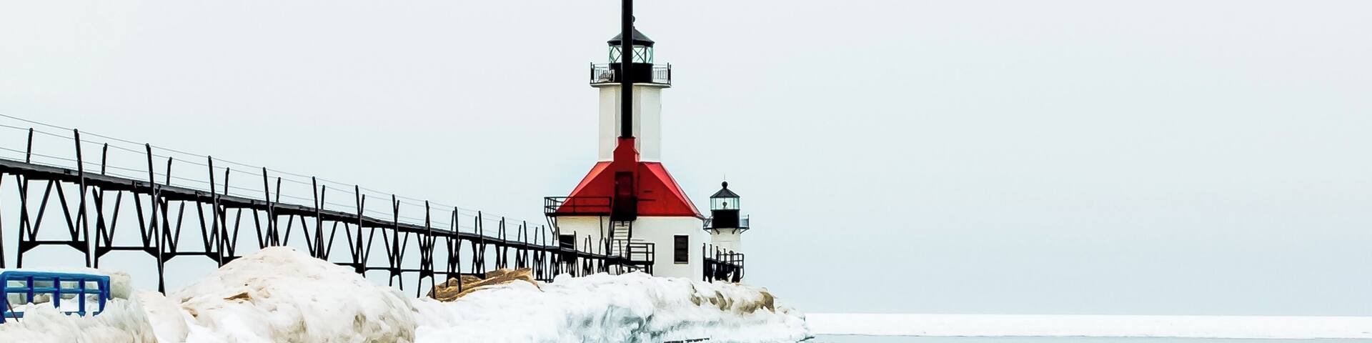A lighthouse with a red roof and a white base, surrounded by ice and snow, with a calm body of water in the foreground.