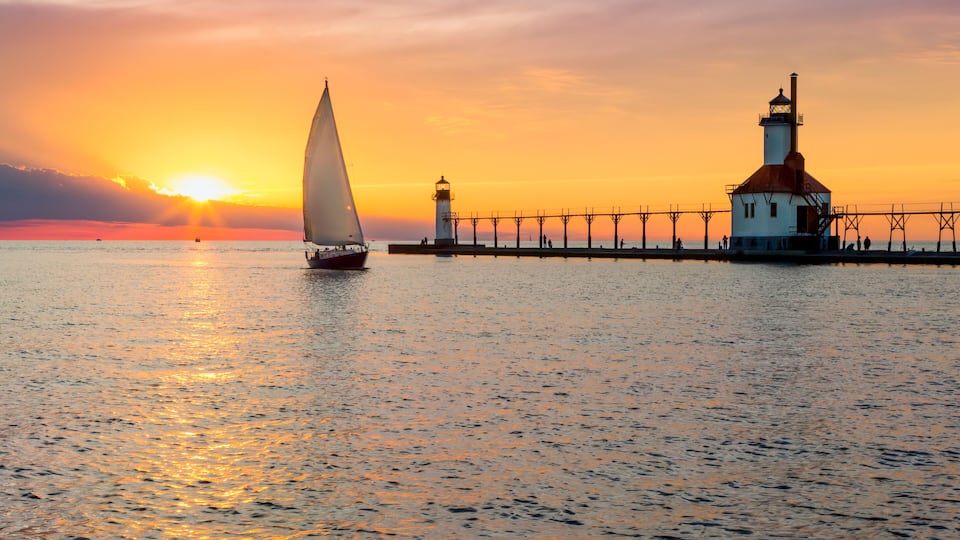 A sailboat on calm waters, a lighthouse, and a pier extending into the sea during sunset.