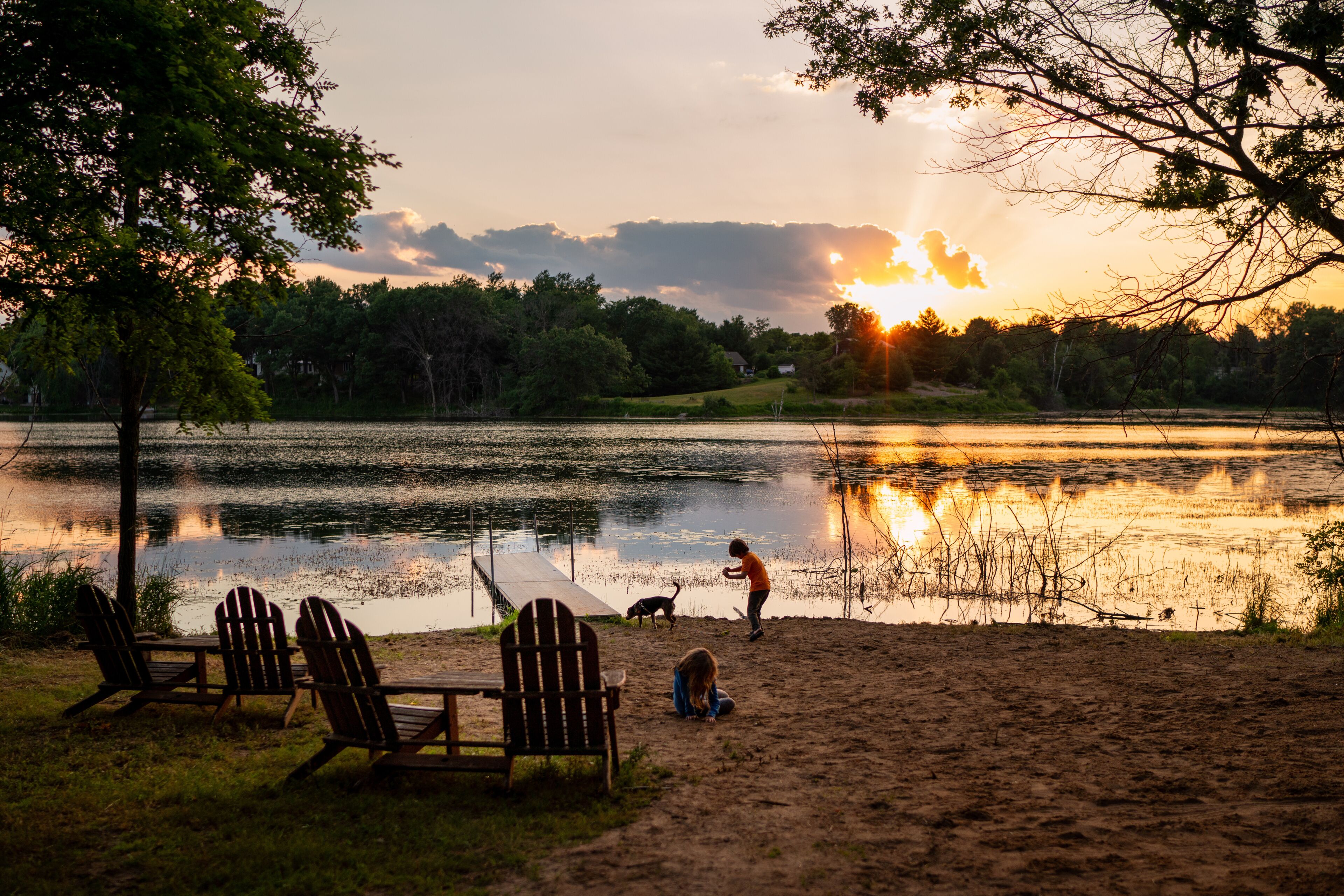 A sunset over a lake with chairs and a dog on the shore.