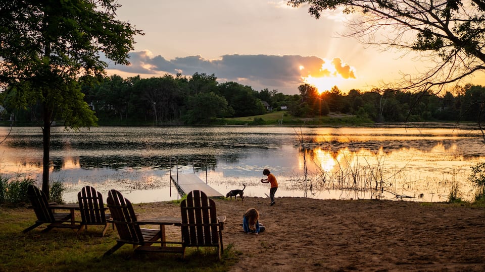 A sunset over a lake with chairs and a dog on the shore.