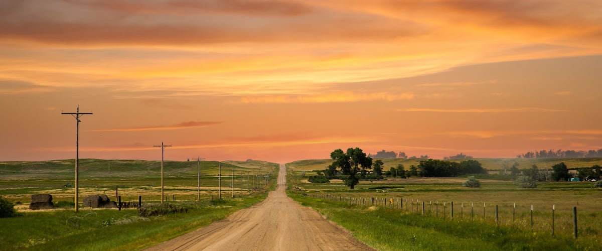 A rural dirt road flanked by fields and a sunset sky.