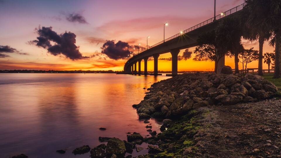 A bridge at sunset with a calm river and rocky shore.