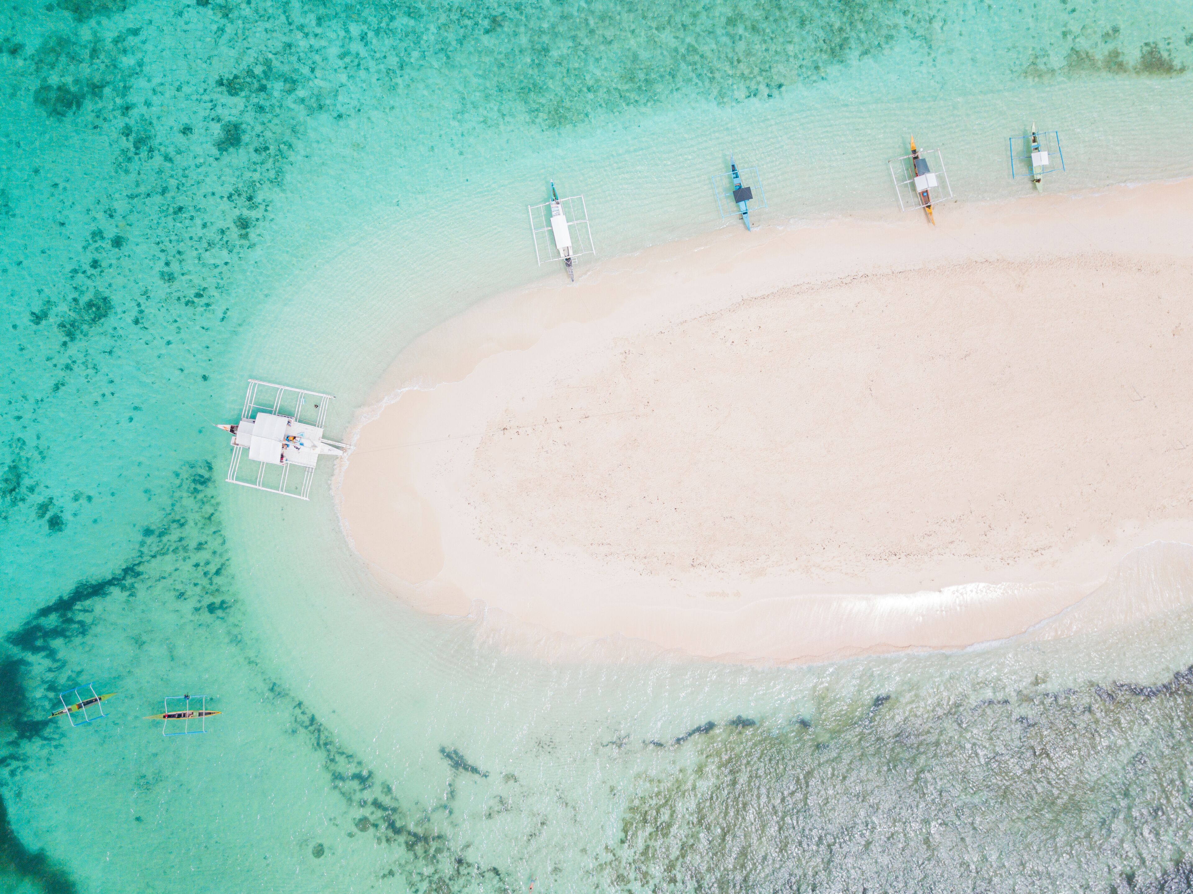 An aerial view of a beach with clear turquoise waters, a sandy shore, and several small boats anchored near the water's edge.