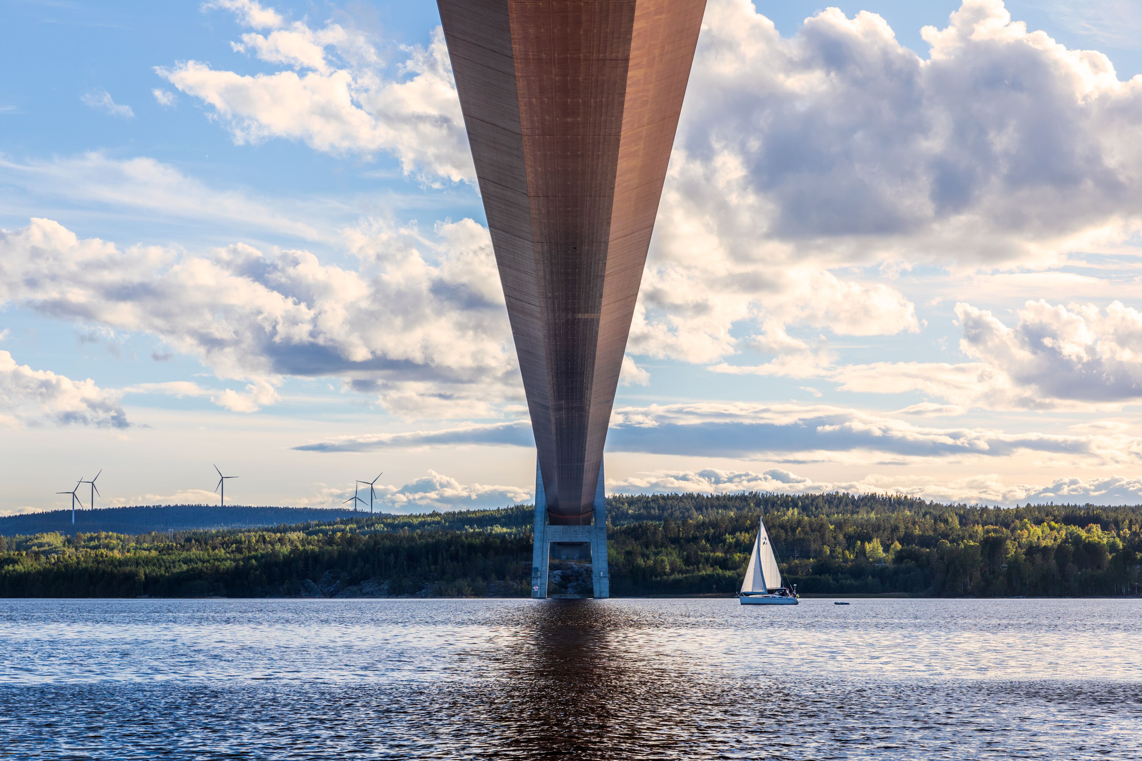 A bridge over water with a sailboat, wind turbines, and a forested hill in the background.