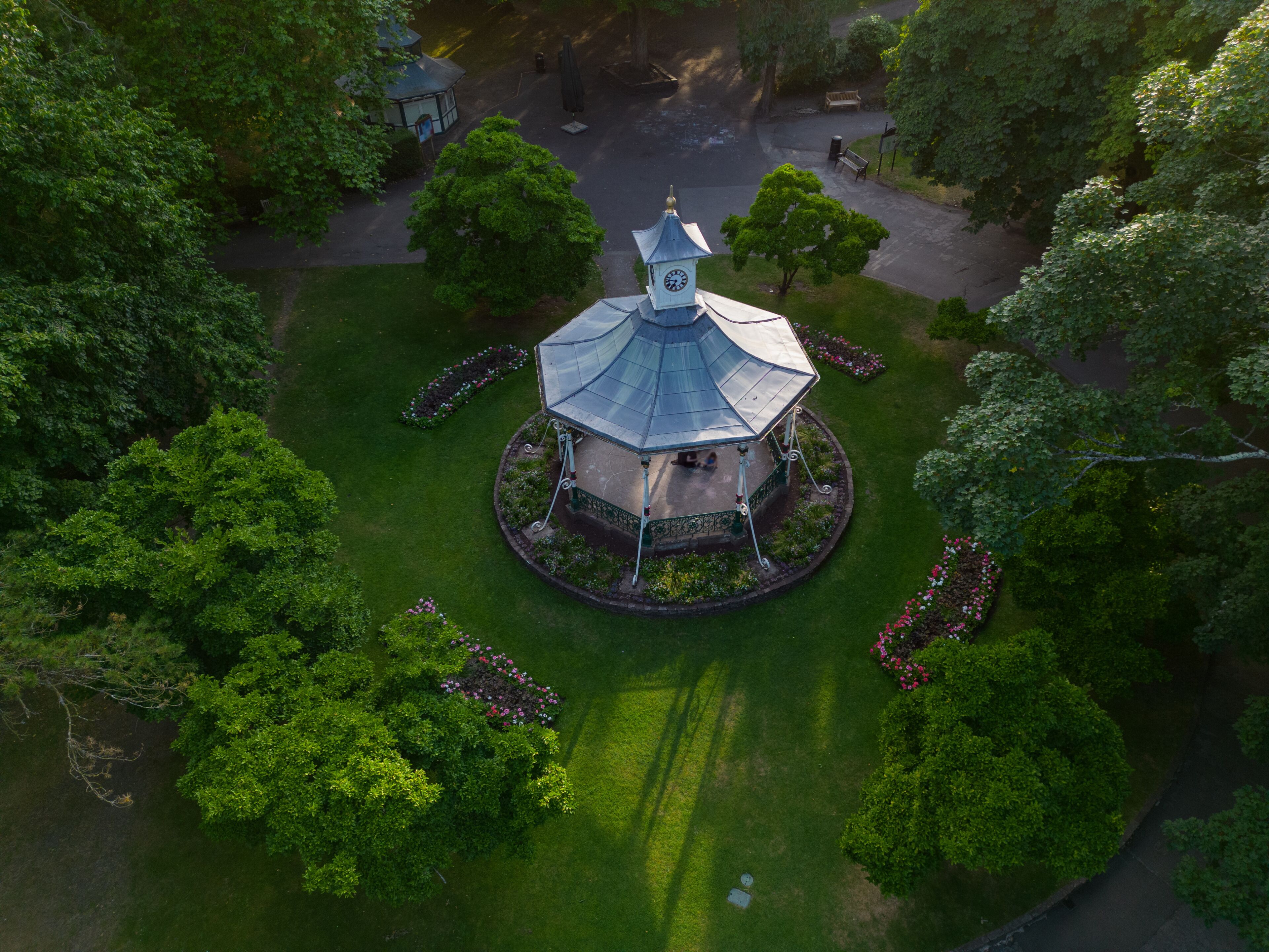 Vista aérea de um gazebo com torre do relógio, cercado por vegetação exuberante e um gramado bem cuidado.