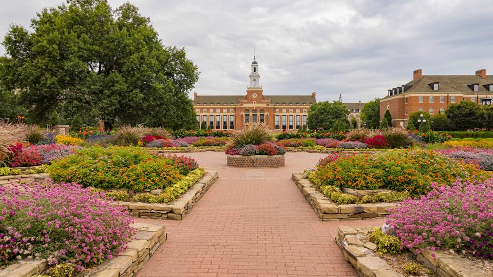A brick pathway leading to a building with a tower, surrounded by a garden with various colorful flowers.