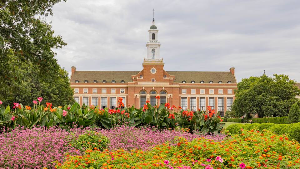 A large brick building with a clock tower, surrounded by colorful flower beds.