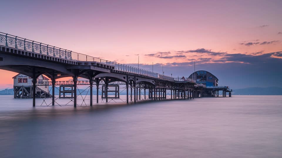 A pier with a lighthouse and a building at the end, extending into calm water.