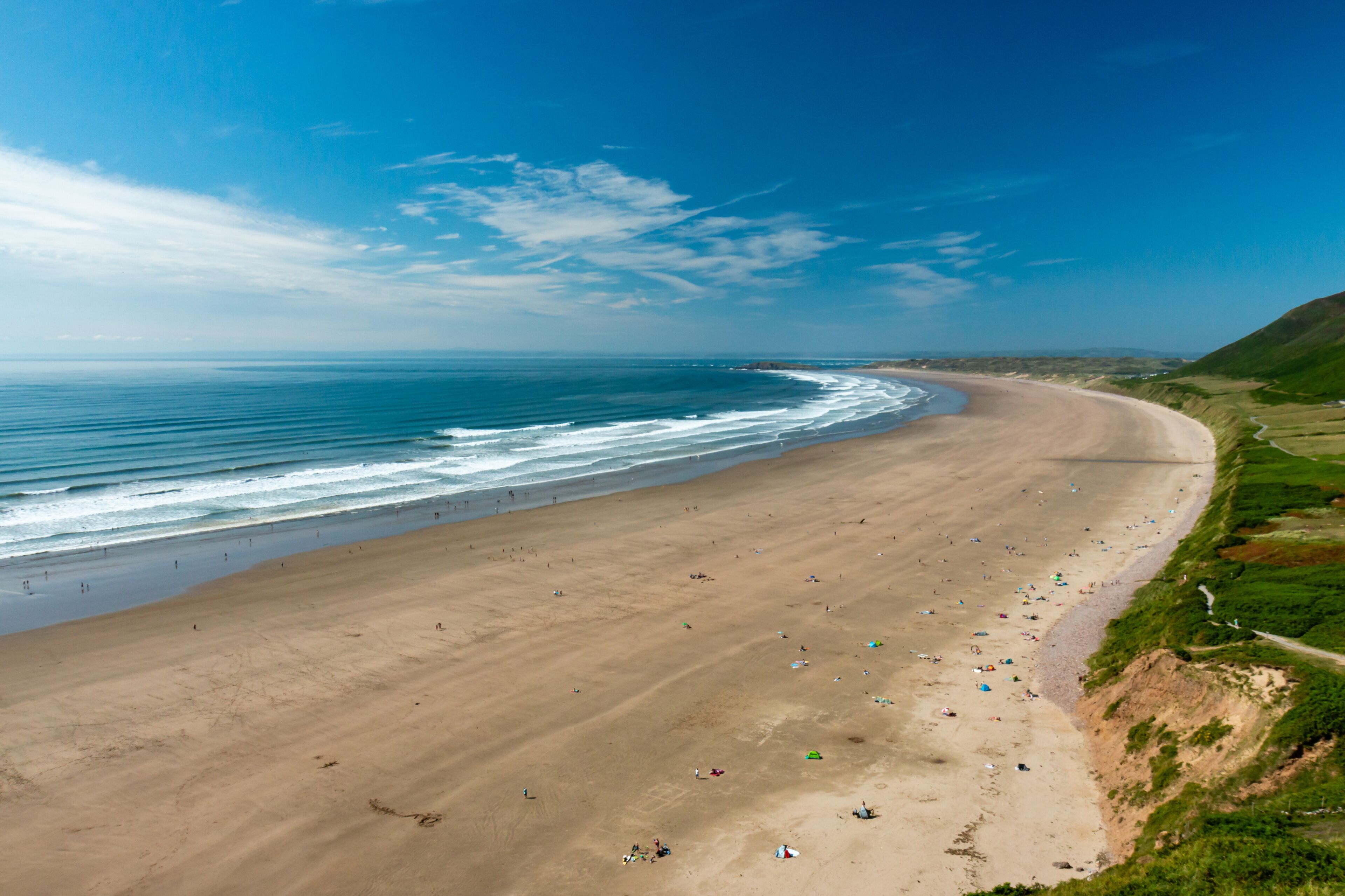 Uma extensa praia de areia com ondas suaves, céu azul límpido e um morro verde ao fundo.