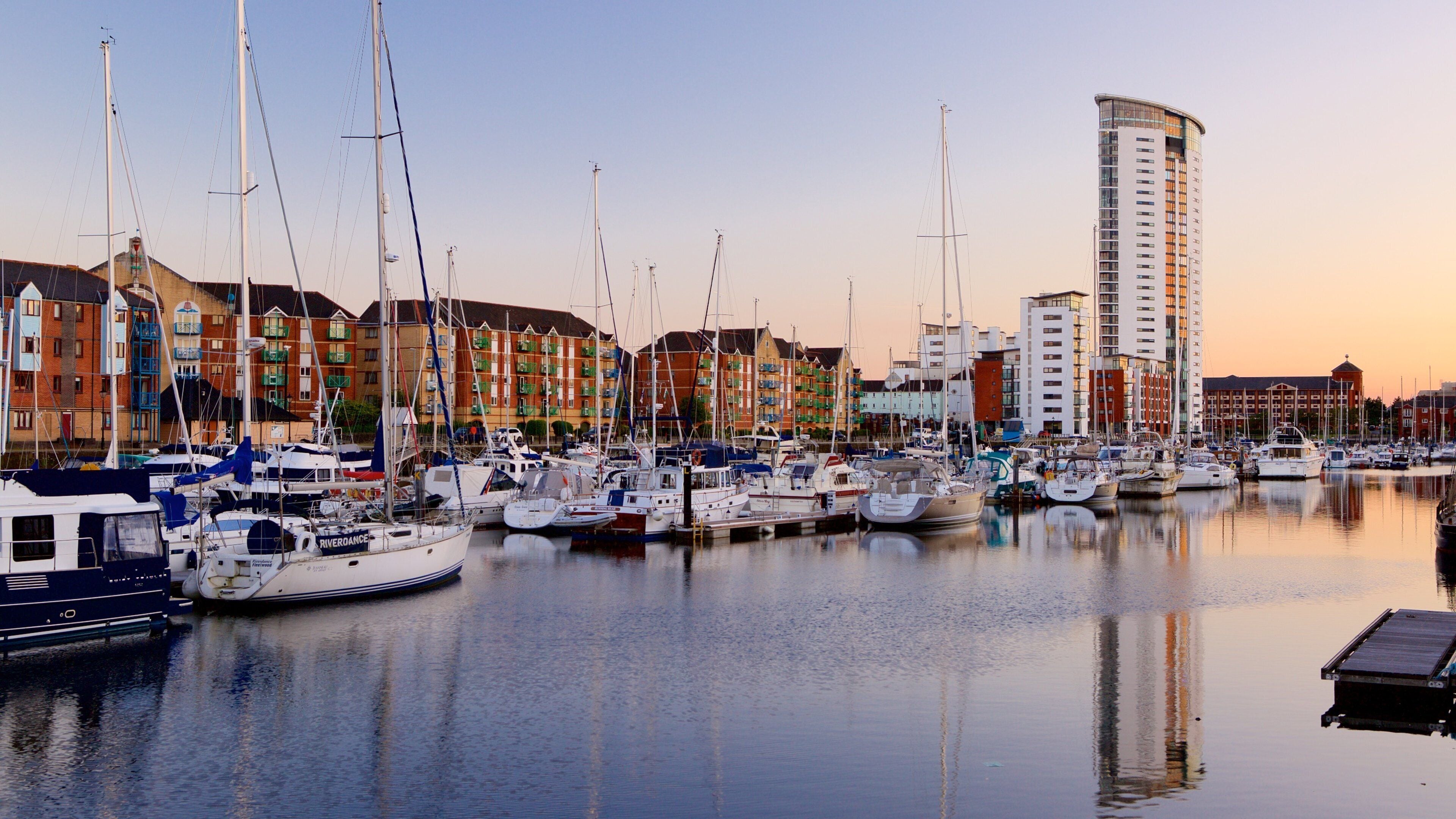 A marina with numerous sailboats docked, surrounded by colorful buildings and a tall modern structure.