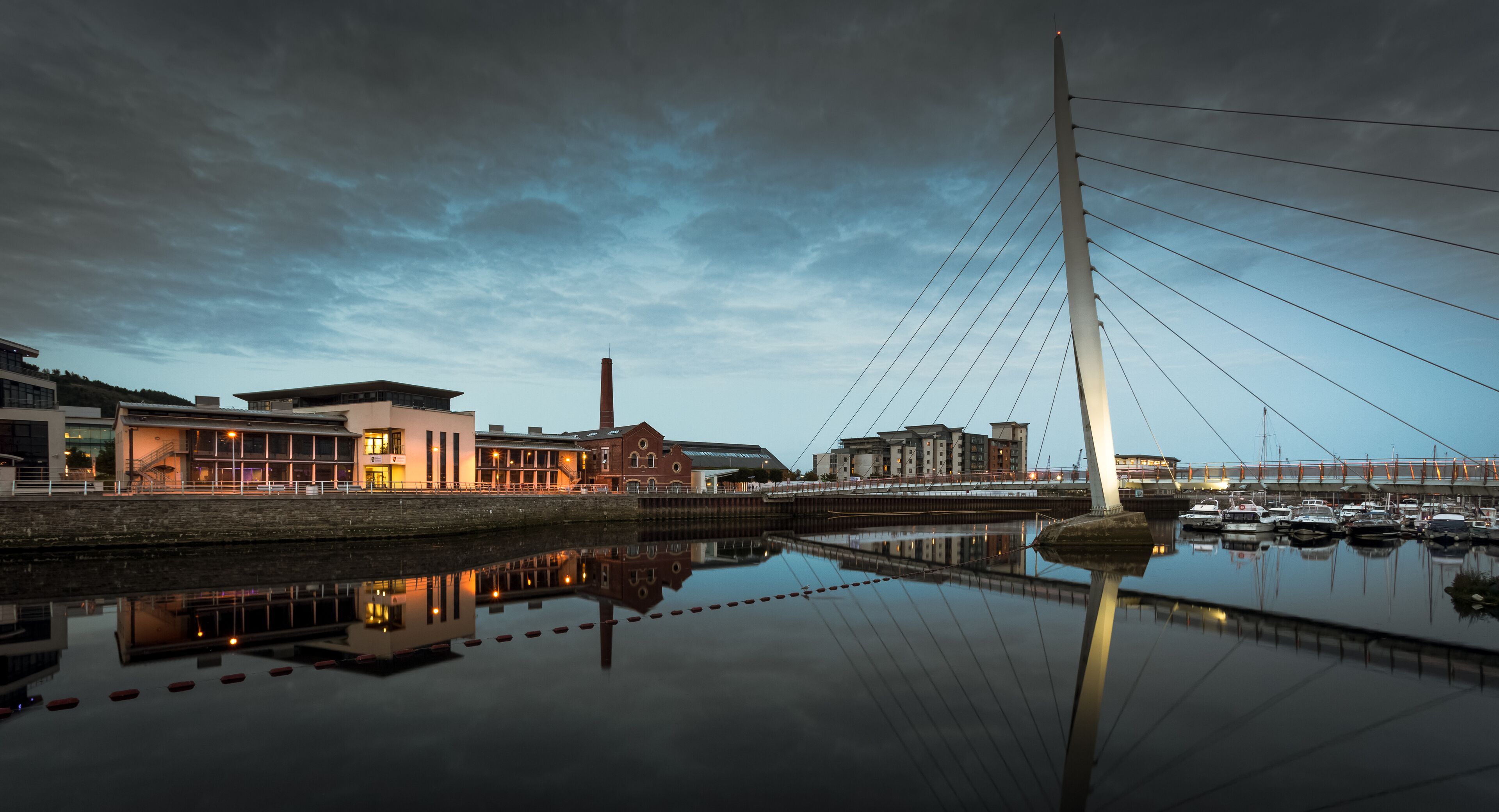 A modern bridge with cables, a calm river reflecting buildings, and a cloudy sky.