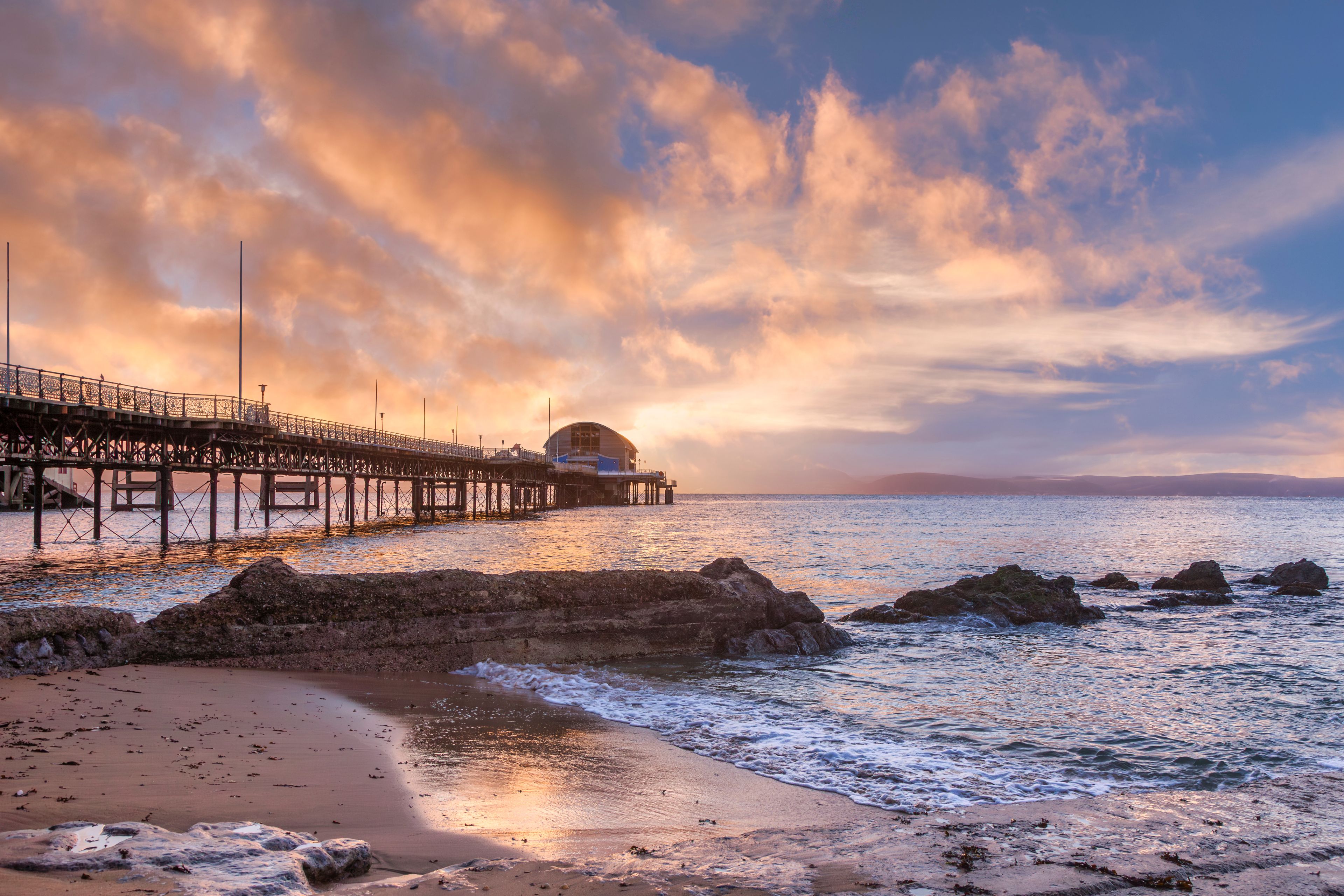 A pier extending into the sea with a building at the end, surrounded by rocky outcrops and a sunset sky.