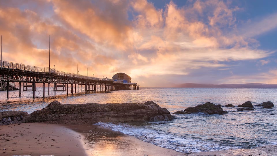 A pier extending into the sea with a building at the end, surrounded by rocky outcrops and a sunset sky.