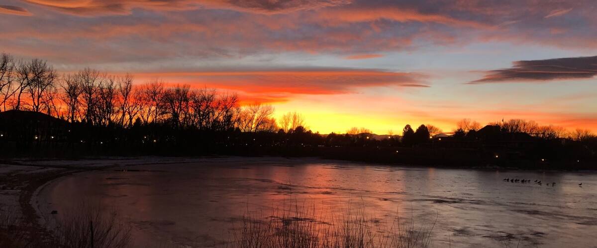 A sunset over a frozen lake with silhouettes of trees and buildings.