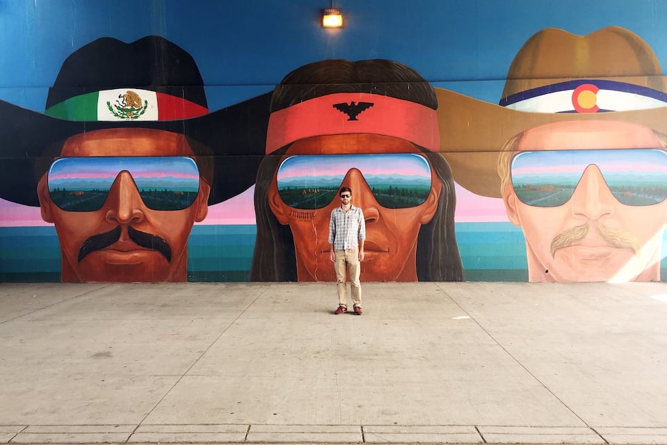 A mural with three large faces wearing different hats, a person standing in front, and a blue sky in the background.