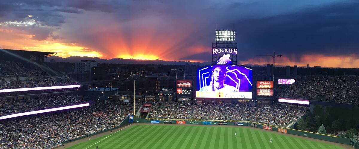 A baseball stadium at sunset with a large crowd, a scoreboard, and advertisements.